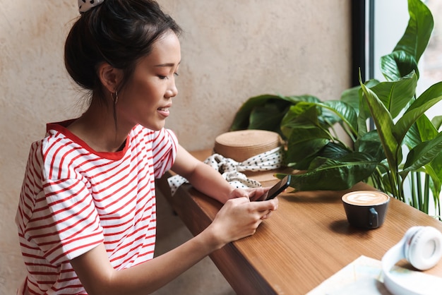Retrato de mujer asiática atractiva sonriendo y sosteniendo el teléfono celular mientras está sentado en el café en el interior