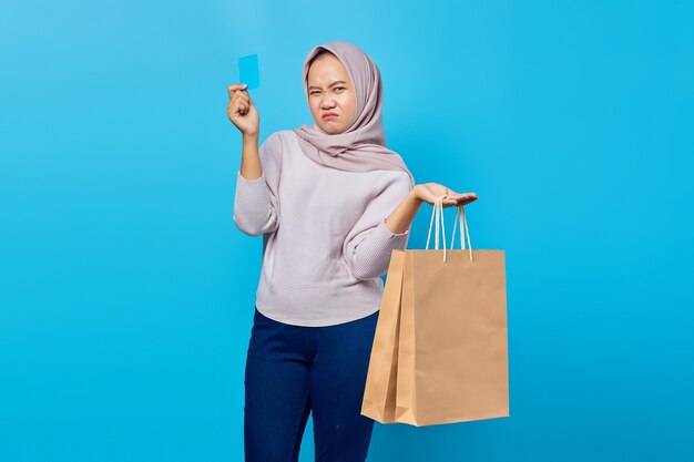 Retrato de mujer asiática atractiva sosteniendo la bolsa de compras y mostrando la tarjeta de crédito sobre fondo azul.