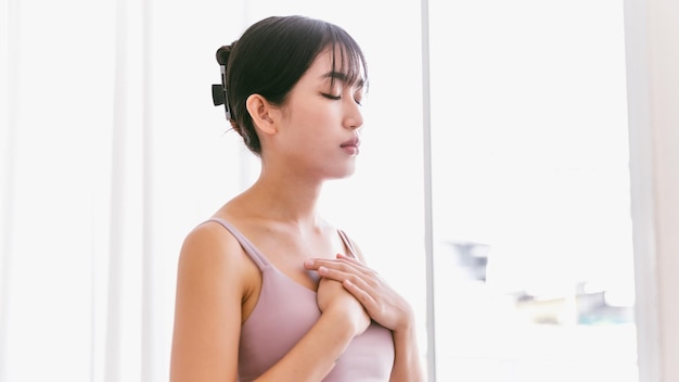 Retrato de una mujer asiática deportiva con las manos en el pecho y cierra los ojos inhala y exhala se reúne con la respiración practicando yoga meditando para la relajación y la atención plena