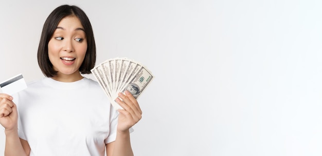 Retrato de mujer asiática con dinero en dólares y tarjeta de crédito que parece impresionada y asombrada de pie...