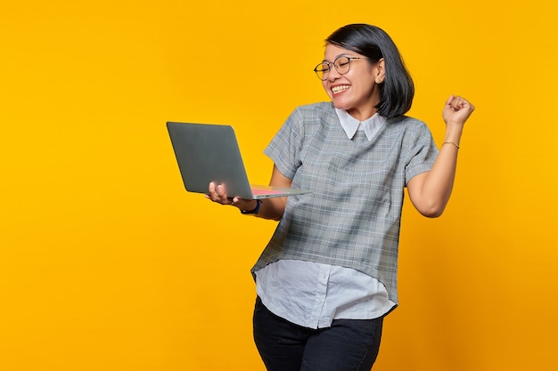 Retrato de mujer asiática emocionada sosteniendo portátil sobre fondo amarillo