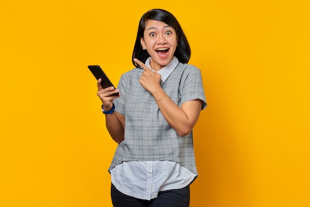 Retrato de mujer asiática emocionada sosteniendo teléfono celular y señalando con el dedo en el espacio vacío sobre fondo amarillo