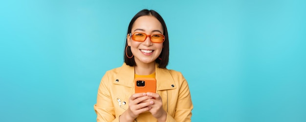 Retrato de mujer asiática entusiasta en gafas de sol usando teléfono móvil sonriendo y riendo mirando h