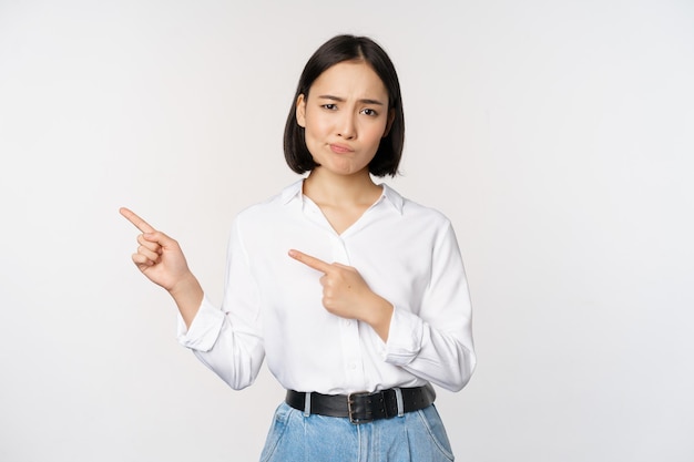 Retrato de una mujer asiática escéptica frunciendo el ceño señalando con el dedo a la izquierda y quejándose expresa disgusto o desaprobación de pie contra el fondo blanco