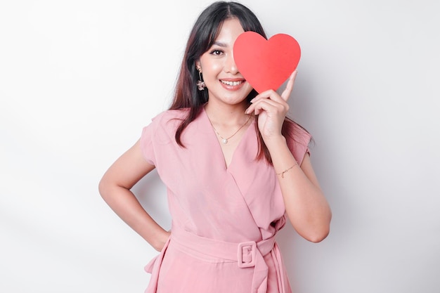 Un retrato de una mujer asiática feliz con una blusa rosa sosteniendo un papel rojo en forma de corazón aislado por un fondo blanco