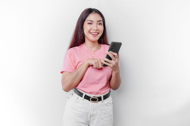 Un retrato de una mujer asiática feliz lleva una camiseta rosa y sostiene su teléfono aislado por un fondo blanco