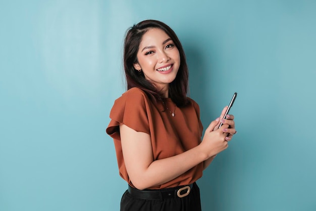 Un retrato de una mujer asiática feliz sonríe y sostiene su teléfono inteligente con una camisa marrón