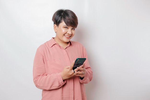 Un retrato de una mujer asiática feliz sonríe y sostiene su teléfono inteligente con una camisa rosa aislada por un fondo blanco