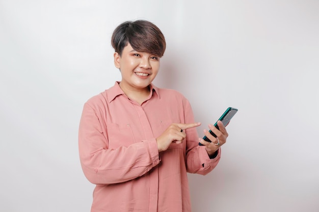 Un retrato de una mujer asiática feliz sonríe y sostiene su teléfono inteligente con una camisa rosa aislada por un fondo blanco