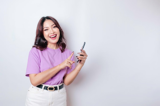 Un retrato de una mujer asiática feliz sonríe y sostiene su teléfono inteligente con una camiseta morada lila aislada por un fondo blanco