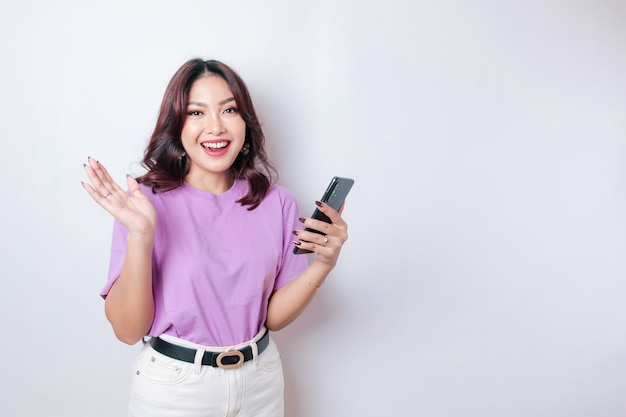 Un retrato de una mujer asiática feliz sonríe y sostiene su teléfono inteligente con una camiseta morada lila aislada por un fondo blanco