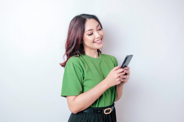 Un retrato de una mujer asiática feliz sonríe y sostiene su teléfono inteligente con una camiseta verde aislada por un fondo blanco