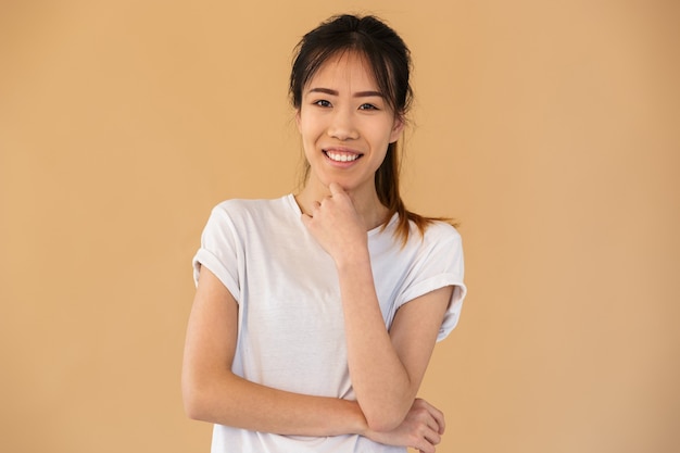 Retrato de mujer asiática feliz vistiendo camiseta básica sonriendo y mirando al frente aislado sobre pared beige en estudio