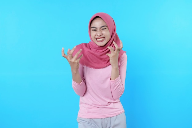 Retrato de mujer asiática gritando enojado aislado sobre fondo azul con hijab y camiseta rosa