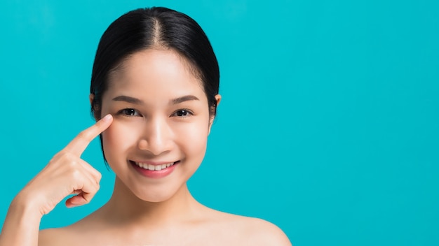 Retrato de la mujer asiática hermosa confiada con la mano al lado del ojo en cara y de la sonrisa aislada en fondo azul del color con el tiro del estudio. Cuidado de la piel para el concepto de rostro saludable.