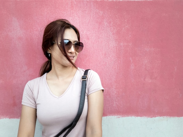 Retrato de la mujer asiática hermosa con las gafas de sol en una ciudad sobre fondo rosado de la pared