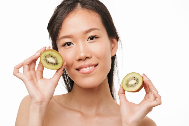 Retrato de una mujer asiática hermosa joven emocionada feliz que presenta aislada sobre la pared blanca que sostiene el kiwi