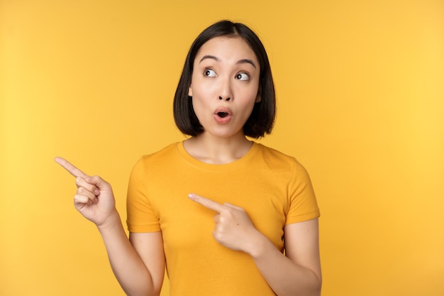 Retrato de mujer asiática intrigada mirando y señalando con el dedo a la izquierda en un anuncio que muestra algo interesante de pie sobre fondo amarillo