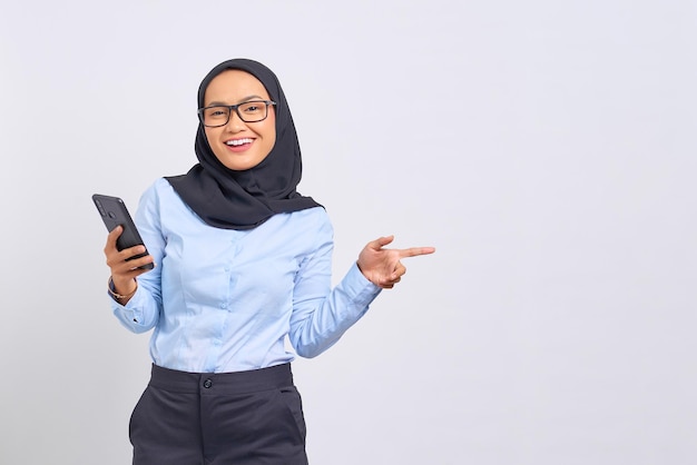 Retrato de mujer asiática joven alegre que sostiene el teléfono móvil y que apunta el dedo lejos en el espacio de la copia aislado en el fondo blanco