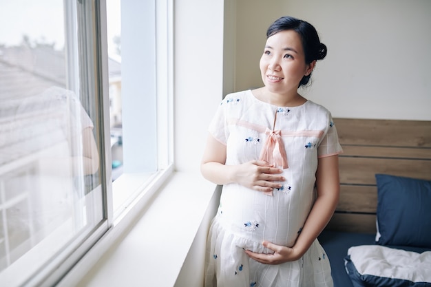 Retrato de mujer asiática joven embarazada alegre de pie en la ventana en el dormitorio y tocando el vientre