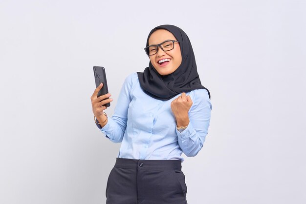 Retrato de mujer asiática joven emocionada celebrando con teléfono móvil aislado sobre fondo blanco.