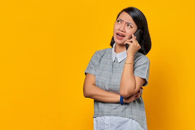 Retrato de mujer asiática joven enojada mientras recibe una llamada entrante en el teléfono inteligente sobre fondo amarillo