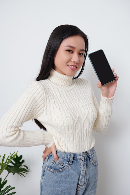 Retrato de mujer asiática joven feliz en suéter blanco cálido mostrando smartphone con pantalla vacía negra