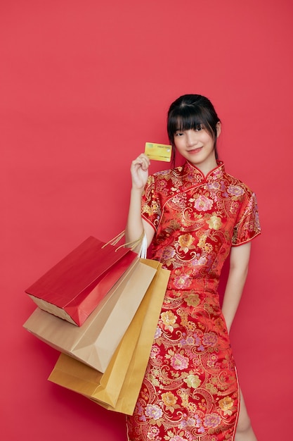 Retrato de mujer asiática joven linda usar vestido chino con una bolsa de compras y tarjeta de crédito para el año nuevo chino en una pared roja