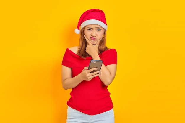 Retrato de mujer asiática joven con sombrero de Navidad pensando y enviando mensajes en el teléfono inteligente