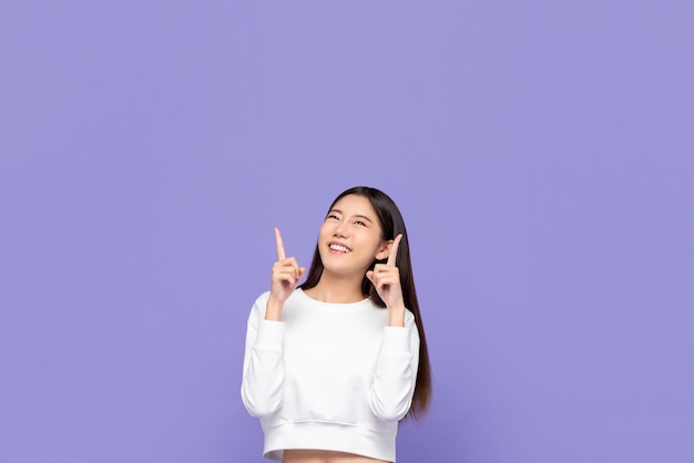 Retrato de mujer asiática joven sonriente linda que señala las manos hacia arriba y mirando copyspace arriba en pared púrpura aislado