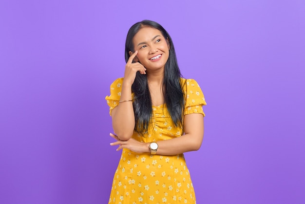 Retrato de mujer asiática joven sonriente pensando en una idea y mirando a otro lado sobre fondo púrpura