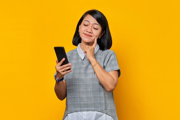 Retrato de mujer asiática joven sonriente pensando en pregunta con el dedo en la barbilla y sosteniendo el teléfono móvil