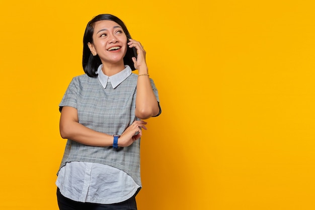 Retrato de mujer asiática joven sonriente que recibe la llamada entrante en el teléfono inteligente y mirando a un lado sobre fondo amarillo