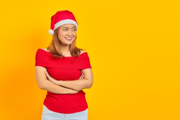 Retrato de mujer asiática joven sonriente con sombrero de navidad cruzando las manos sobre fondo amarillo