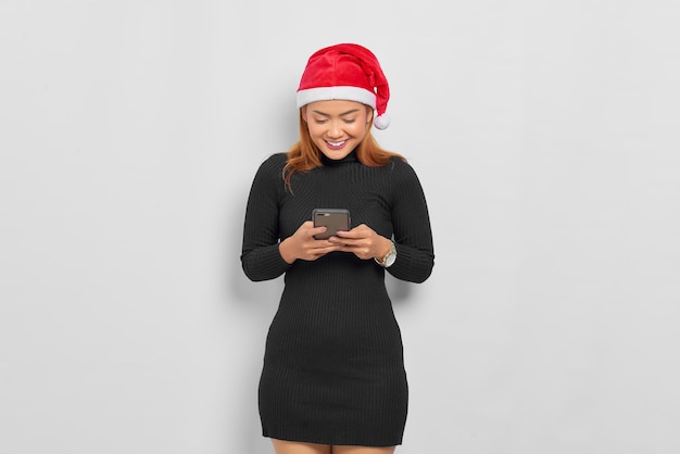 Retrato de mujer asiática joven sonriente con sombrero de Santa Claus mediante teléfono móvil aislado sobre fondo blanco.