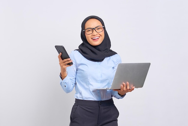 Retrato de mujer asiática joven sonriente sosteniendo un portátil y un teléfono móvil aislado sobre fondo blanco.