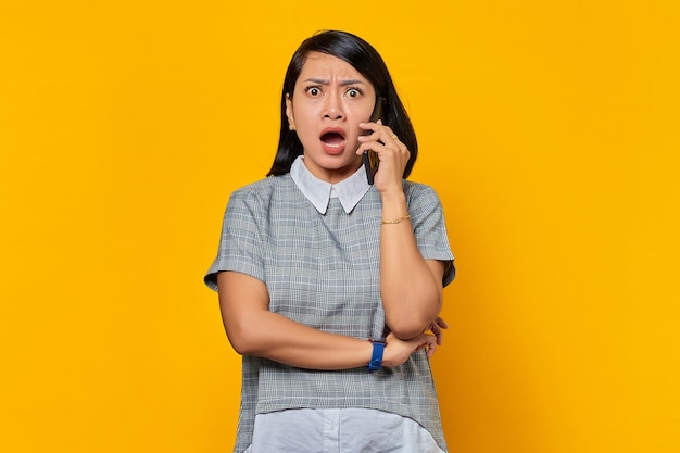 Retrato de mujer asiática joven sorprendida mientras recibe una llamada entrante en el teléfono inteligente sobre fondo amarillo