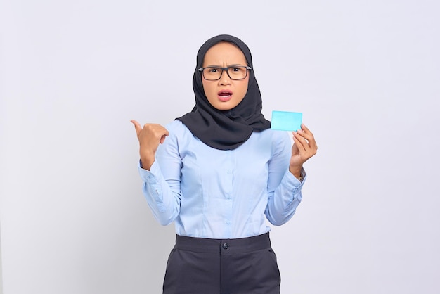 Retrato de mujer asiática joven sorprendida que sostiene la tarjeta de crédito y que señala el pulgar hacia arriba en el espacio de la copia aislado en el fondo blanco