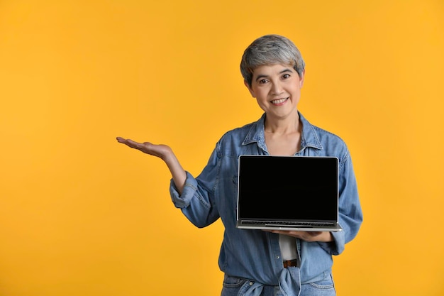 Retrato de una mujer asiática de mediana edad de 50 años con una camisa de mezclilla casual camiseta blanca sosteniendo una computadora portátil con una pantalla en blanco y una palma de la mano abierta aislada en un fondo amarillo