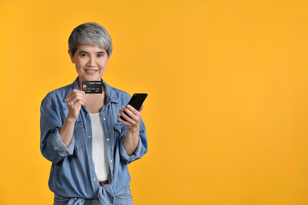 Retrato de una mujer asiática de mediana edad de 50 años con pantalones de mezclilla casual camiseta blanca con teléfono inteligente y compras con tarjeta de crédito en línea aisladas en un fondo de color mirando y sonriendo a la cámara