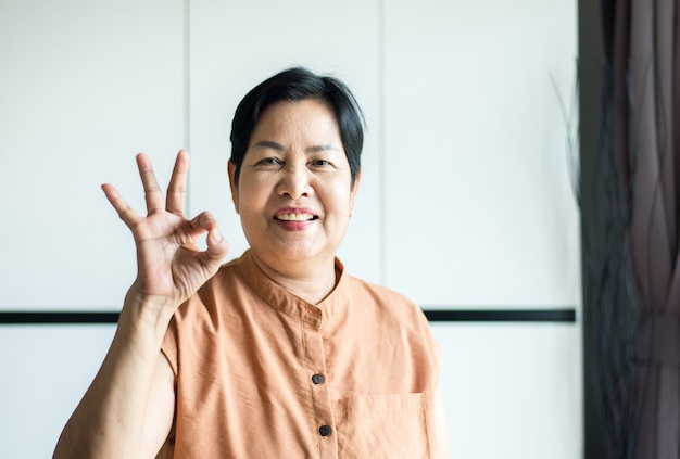 Retrato de mujer asiática de mediana edad con rostro sonriente y mostrando el signo de ok en casa, concepto de seguro médico