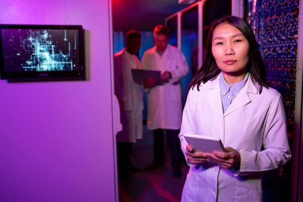 Retrato de mujer asiática moderna de contenido en bata de laboratorio sosteniendo tableta digital y trabajando en la sala de servidores con luz de neón