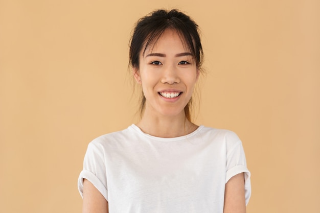 Retrato de mujer asiática positiva vistiendo camiseta básica sonriendo y mirando al frente aislado sobre pared beige en estudio