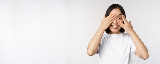 Retrato de mujer asiática que cubre los ojos esperando sorpresa con los ojos vendados sonriendo y mirando a la cámara de pie sobre fondo blanco.