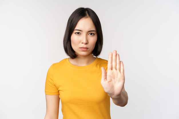 Retrato de una mujer asiática que parece seria y enojada mostrando dejar de prohibir el gesto tabú que prohíbe algo de pie en una camiseta amarilla sobre fondo blanco