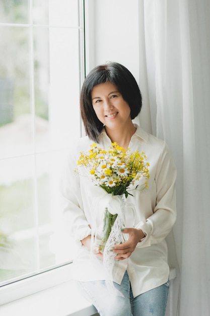 Retrato de una mujer asiática con un ramo de flores silvestres en casa cerca de la ventana
