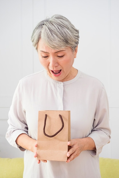 retrato de una mujer asiática senior una bolsa de regalo, sorprendida en casa.