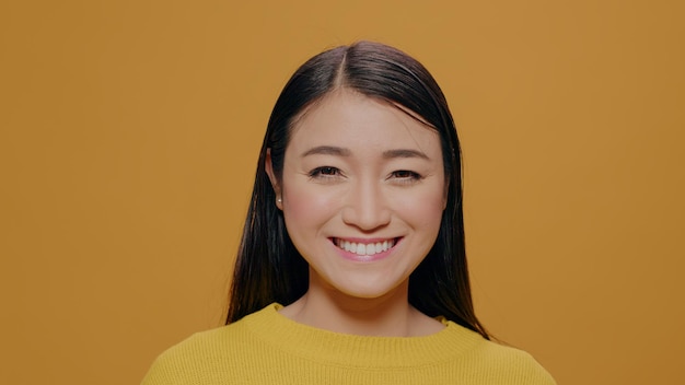 Retrato de mujer asiática sonriendo frente a cámara, de pie sobre fondo amarillo. Persona positiva y casual mirando a cámara, con expresión facial natural. Adulto agraciado mostrando sonrisa