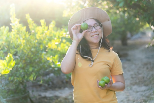 Retrato de una mujer asiática sonriendo usando un sombrero cosechando limones verdes en el jardín de la huerta de la granja Estilo de vida campesino mirando la cámara al aire libre Concepto de granja de limón orgánico y agricultor