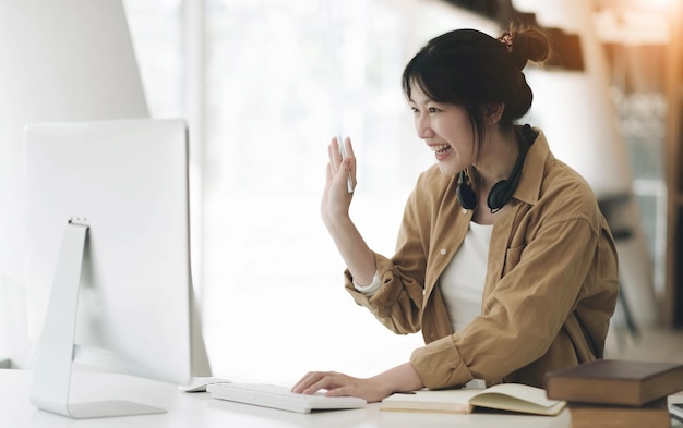 Retrato de una mujer asiática sonriente con auriculares posando para una foto en el lugar de trabajo mujer feliz y emocionada con auriculares mirando una computadora portátil sentada en el escritorio con una computadora portátil haciendo videollamadasxA